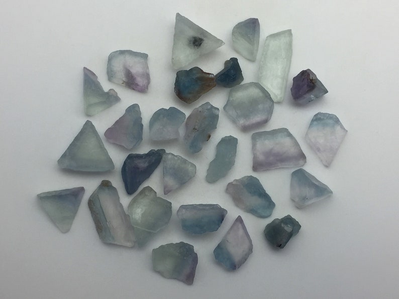 May include: A collection of 25 blue and purple fluorite crystals of various shapes and sizes. The crystals are translucent and have a slight shimmer.