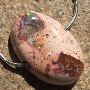 Op de afbeelding: Een zilveren ketting met een hanger gemaakt van een lichtbruine, ruwe steen met iriserende groene en oranje opaal-insluitsels.