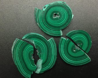 May include: Four pieces of broken malachite stone, each with a swirling green pattern. The pieces are arranged on a dark gray surface.