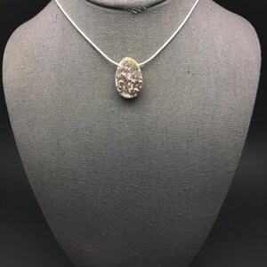 May include: A silver necklace featuring a pendant with a speckled, oval-shaped design. The pendant has a mix of brown, white, and yellow tones. The necklace is displayed on a gray mannequin.