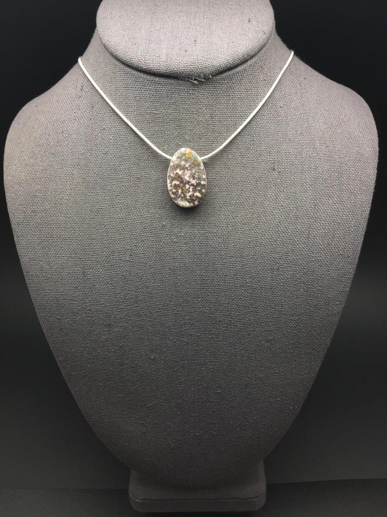 May include: A silver necklace featuring a pendant with a speckled, oval-shaped design. The pendant has a mix of brown, white, and yellow tones. The necklace is displayed on a gray mannequin.