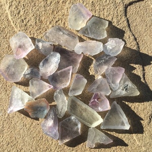 May include: A collection of 20+ clear, purple, and green fluorite crystals on a sandy surface. The crystals are various sizes and shapes, some are triangular, some are rectangular, and some are irregular.
