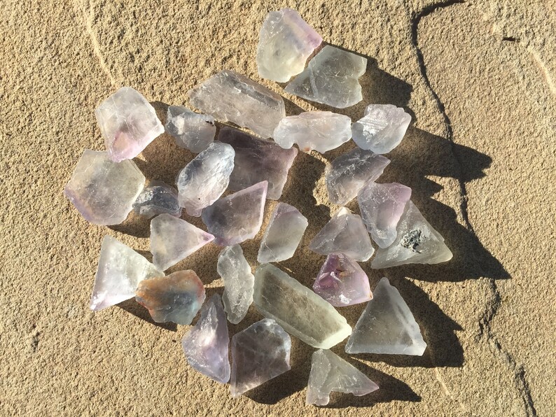 May include: A collection of 20+ clear, purple, and green fluorite crystals on a sandy surface. The crystals are various sizes and shapes, some are triangular, some are rectangular, and some are irregular.
