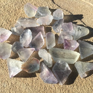 May include: A collection of purple and green fluorite crystals, some with a slight iridescent sheen, scattered on a sandy surface.
