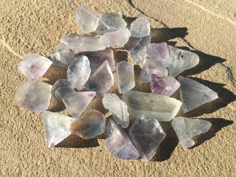 May include: A collection of purple and green fluorite crystals, some with a slight iridescent sheen, scattered on a sandy surface.