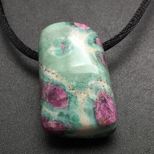 May include: A green and pink stone pendant on a black cord. The polished stone has a rectangular shape with rounded edges and a mix of green, pink, and white inclusions. The cord is a woven black material.