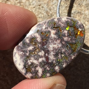 May include: A polished, oval-shaped pendant with a unique pattern. The stone features a mix of colors, including white, purple, green, and orange, with a speckled texture. It is suspended from a silver chain, creating a striking piece of jewelry.