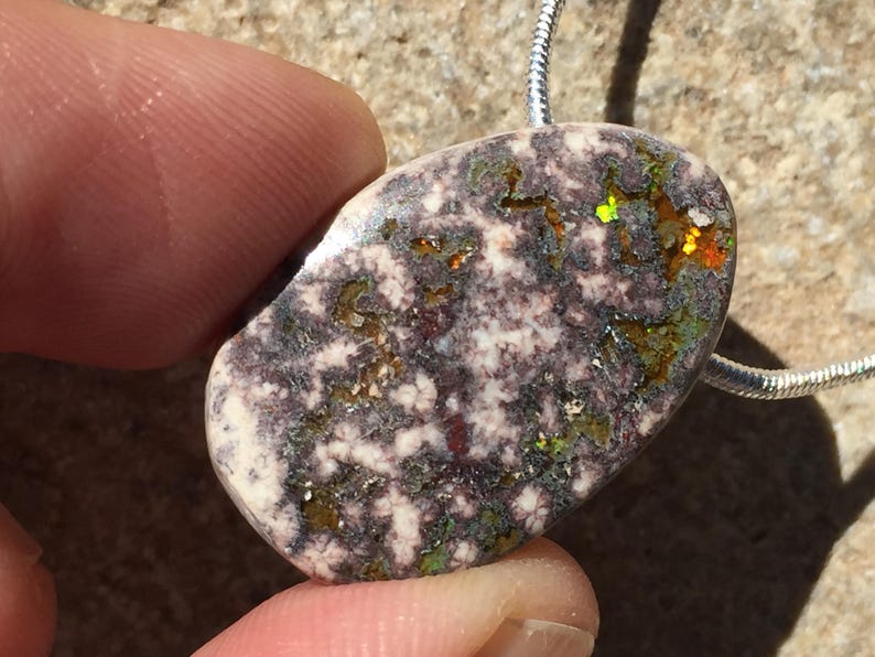 May include: A polished, oval-shaped pendant with a unique pattern. The stone features a mix of colors, including white, purple, green, and orange, with a speckled texture. It is suspended from a silver chain, creating a striking piece of jewelry.