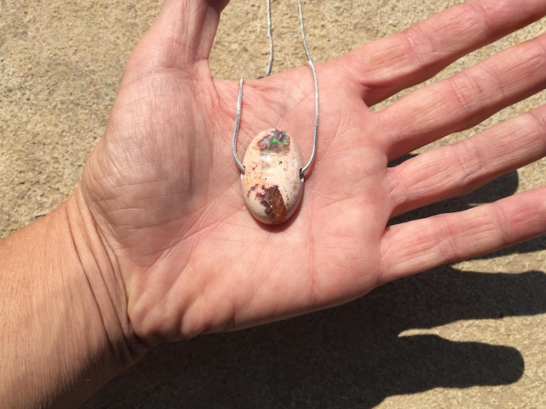 Op de afbeelding: Een zilveren ketting met een lichtroze ovale opaal hanger. De opaal heeft bruine en groene insluitsels.