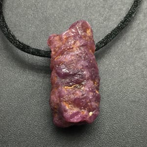 May include: A raw, unpolished ruby pendant with a deep magenta and purple hue, featuring a textured, crystalline surface. The stone is suspended from a thin, black cord, showcasing its natural, irregular shape and multifaceted, rugged appearance.