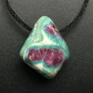 May include: A pendant featuring a polished, irregularly shaped stone with a smooth surface. The stone displays a marbled pattern of vibrant magenta-pink inclusions set against a background of teal, green, and white. It hangs from a black cord.