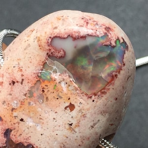 Op de afbeelding: Een roze en witte opaal hanger met een zilveren ketting. De opaal heeft een ruw, getextureerd oppervlak en een grote iriserende vlek.
