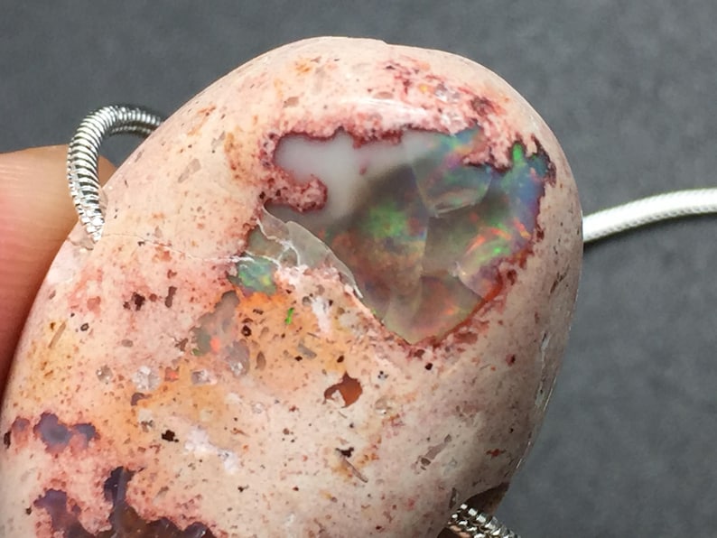 Op de afbeelding: Een roze en witte opaal hanger met een zilveren ketting. De opaal heeft een ruw, getextureerd oppervlak en een grote iriserende vlek.