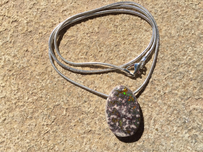 May include: A silver necklace with a pendant. The pendant is an oval-shaped stone with a speckled pattern of white, gray, and black, with flecks of green, orange, and yellow. The necklace is on a textured, tan surface.