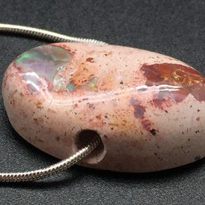 Op de afbeelding: Een roze en rode opaal hanger met een zilveren ketting. De opaal heeft een gladde, ronde vorm en een gat dat erdoor geboord is voor de ketting. De opaal heeft een natuurlijke, ruwe textuur en een verscheidenheid aan kleuren, waaronder roze, rood en wit.
