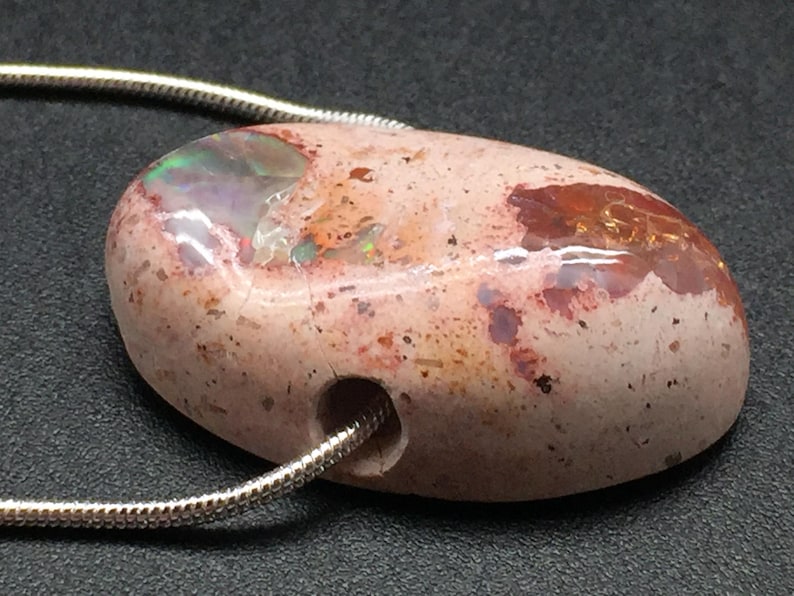 Op de afbeelding: Een roze en rode opaal hanger met een zilveren ketting. De opaal heeft een gladde, ronde vorm en een gat dat erdoor geboord is voor de ketting. De opaal heeft een natuurlijke, ruwe textuur en een verscheidenheid aan kleuren, waaronder roze, rood en wit.