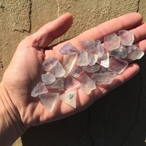 May include: A hand holding a collection of small, clear, purple-hued crystals. The crystals are various shapes and sizes, with some having a more opaque appearance.