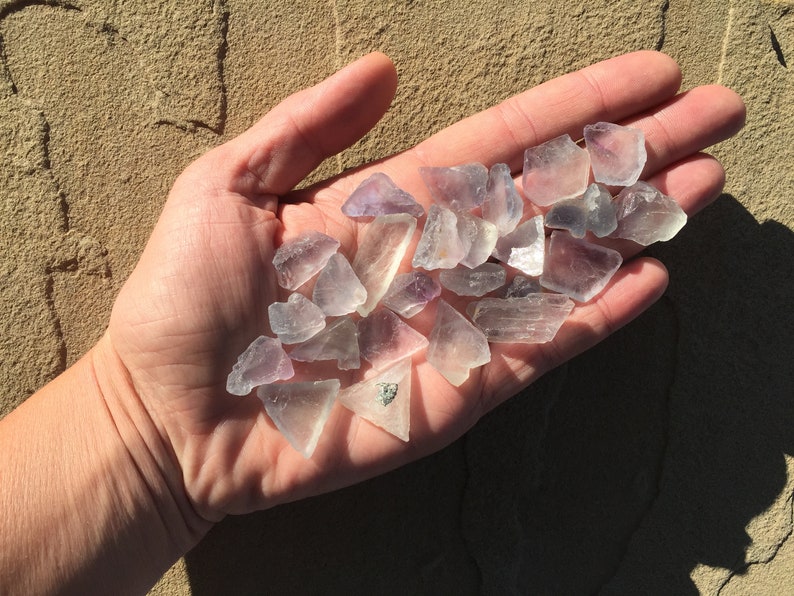 May include: A hand holding a collection of small, clear, purple-hued crystals. The crystals are various shapes and sizes, with some having a more opaque appearance.