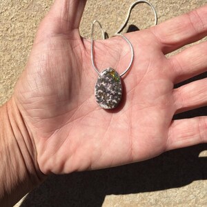 May include: Oval pendant necklace with a silver chain. The pendant is a speckled mix of white, gray, and black with hints of green and gold. The necklace is displayed on a hand.
