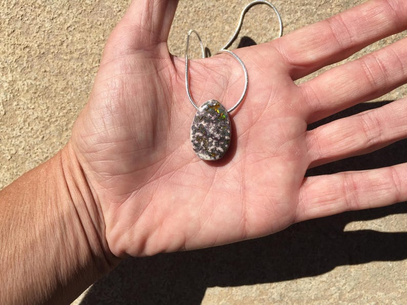 May include: Oval pendant necklace with a silver chain. The pendant is a speckled mix of white, gray, and black with hints of green and gold. The necklace is displayed on a hand.