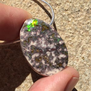 May include: A pendant necklace featuring a polished, oval-shaped stone with a speckled pattern of white, gray, and brown. The stone has iridescent green and yellow highlights. The pendant is attached to a silver chain.