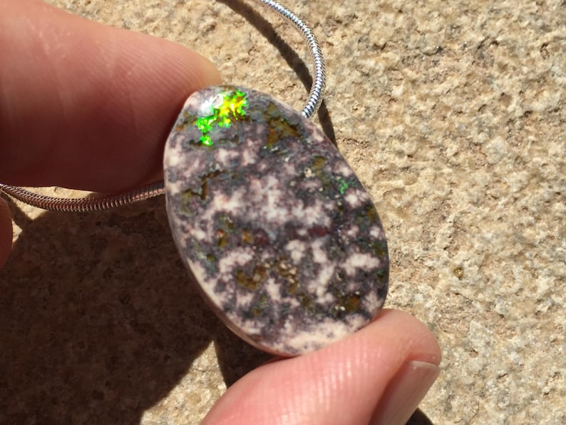 May include: A pendant necklace featuring a polished, oval-shaped stone with a speckled pattern of white, gray, and brown. The stone has iridescent green and yellow highlights. The pendant is attached to a silver chain.