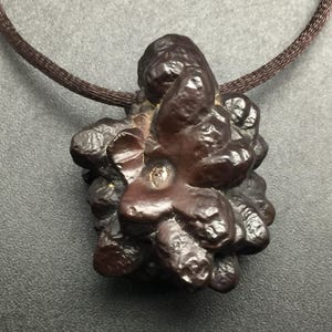 May include: A dark brown pendant necklace with a textured, organic-looking stone. The stone is attached to a brown cord. The pendant has a unique, irregular shape and a small, shiny accent.