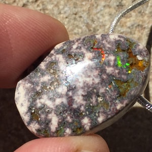 May include: A polished, oval-shaped pendant with a speckled, multi-colored surface. The stone features a mix of white, gray, and brown hues, with flashes of red, green, and yellow. A silver chain is attached to the pendant.