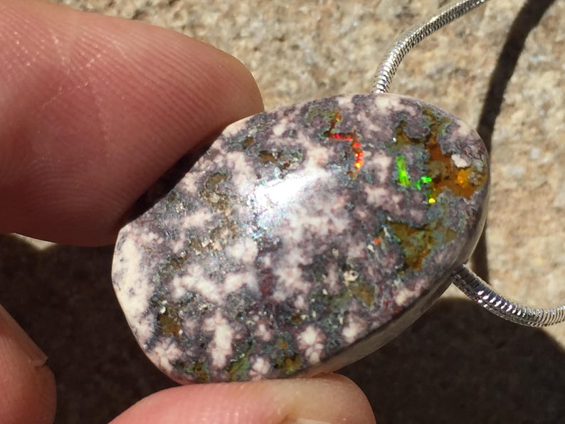May include: A polished, oval-shaped pendant with a speckled, multi-colored surface. The stone features a mix of white, gray, and brown hues, with flashes of red, green, and yellow. A silver chain is attached to the pendant.
