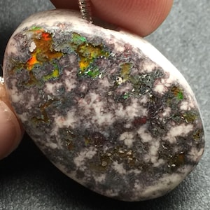 May include: A natural stone pendant with a silver chain. The oval-shaped stone features a mix of colors, including white, gray, orange, green, and yellow. The stone has a textured surface and is likely a type of opal.