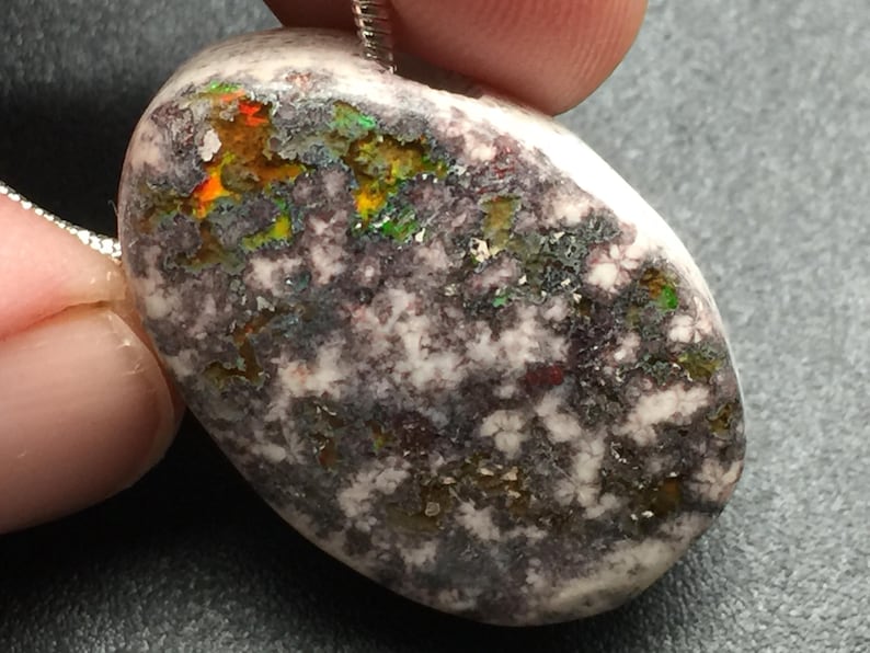 May include: A natural stone pendant with a silver chain. The oval-shaped stone features a mix of colors, including white, gray, orange, green, and yellow. The stone has a textured surface and is likely a type of opal.