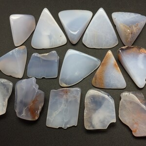 May include: A collection of 16 blue chalcedony stones, some with brown inclusions, in various shapes and sizes. The stones are arranged on a dark grey surface.