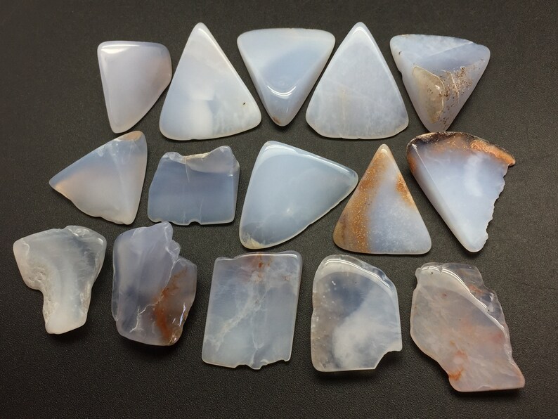 May include: A collection of 16 blue chalcedony stones, some with brown inclusions, in various shapes and sizes. The stones are arranged on a dark grey surface.