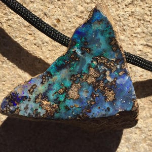 May include: A triangular opal pendant with a black cord. The opal features a vibrant display of blues, greens, and gold, with a rough, natural edge. The stone is set against a neutral background.
