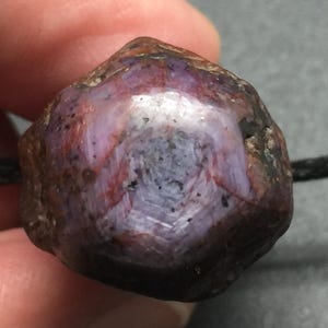 May include: A raw, hexagonal star ruby gemstone pendant strung on a thin black cord. The stone features a deep purple and reddish-brown color palette with a visible six-rayed star pattern across its polished, multifaceted surface.