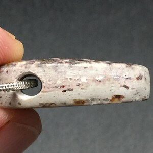 May include: A rectangular pendant with a hole for a necklace. The pendant is off-white with reddish-brown and gray speckles. A silver chain is threaded through the hole. The pendant is held by a person's fingers.