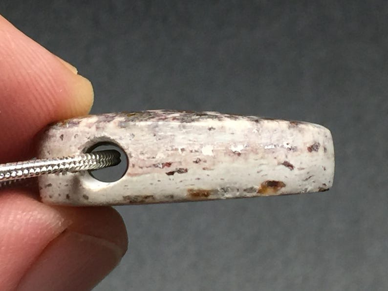 May include: A rectangular pendant with a hole for a necklace. The pendant is off-white with reddish-brown and gray speckles. A silver chain is threaded through the hole. The pendant is held by a person's fingers.
