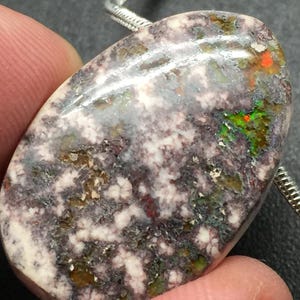 May include: Oval-shaped gemstone pendant with a unique pattern. The stone features a mix of colors, including white, purple, green, and orange. A silver chain is attached to the pendant. The pendant is held by a finger.
