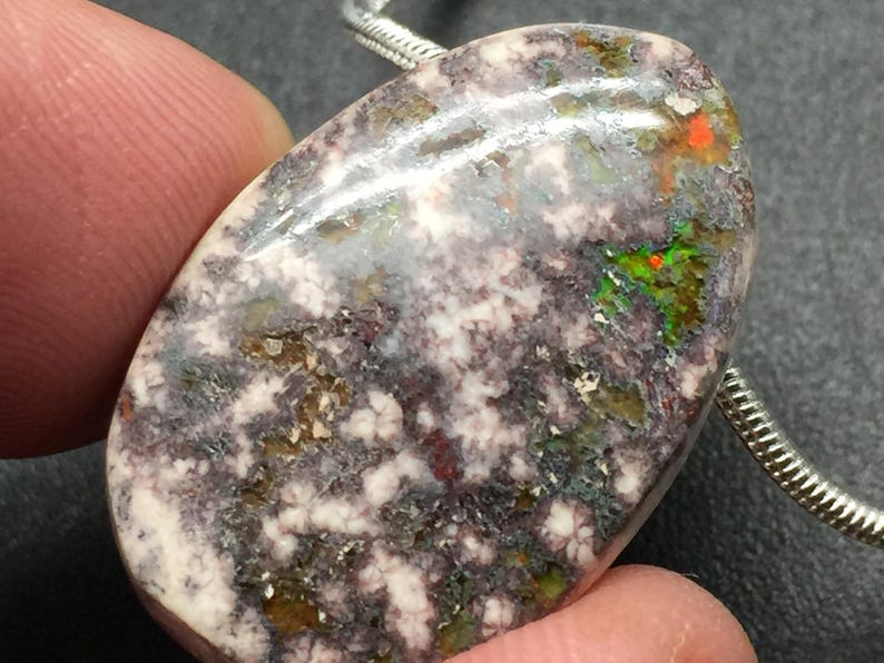 May include: Oval-shaped gemstone pendant with a unique pattern. The stone features a mix of colors, including white, purple, green, and orange. A silver chain is attached to the pendant. The pendant is held by a finger.