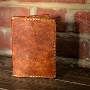 May include: A vertical, rectangular leather passport holder in a medium brown tone with a mottled, textured surface. The item features visible, light-colored stitching along the edges, creating a clean, defined border around the entire structure.