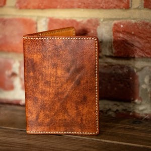 May include: A vertical bi-fold wallet crafted from distressed brown leather with a mottled texture. The wallet features visible light-colored stitching along the edges and a smooth, lighter-toned interior lining visible at the top fold.