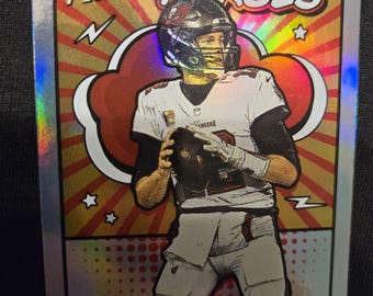 May include: A 2021 Prestige Heroes trading card featuring Tom Brady of the Buccaneers. The card has a silver border and a colorful background with the word "Heroes" in white. Brady is depicted in his uniform, holding a football.