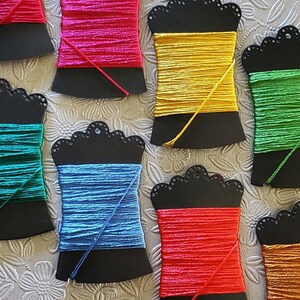 May include: An assortment of colorful embroidery floss spools. Each spool is a different color, including red, pink, yellow, green, blue, and orange. The spools are mounted on black, decorative holders.