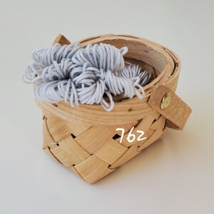 May include: A small, woven basket with a light gray yarn ball inside. The basket has a leather handle with a gold metal clasp and the number "762" written on the side.