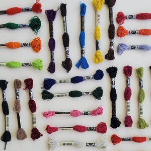 Puede incluir: Surtido de carretes de hilo de bordar en varios colores, incluyendo rojo, verde, azul, amarillo, morado, marr&oacute;n y gris. Los carretes est&aacute;n dispuestos en filas sobre una superficie blanca.