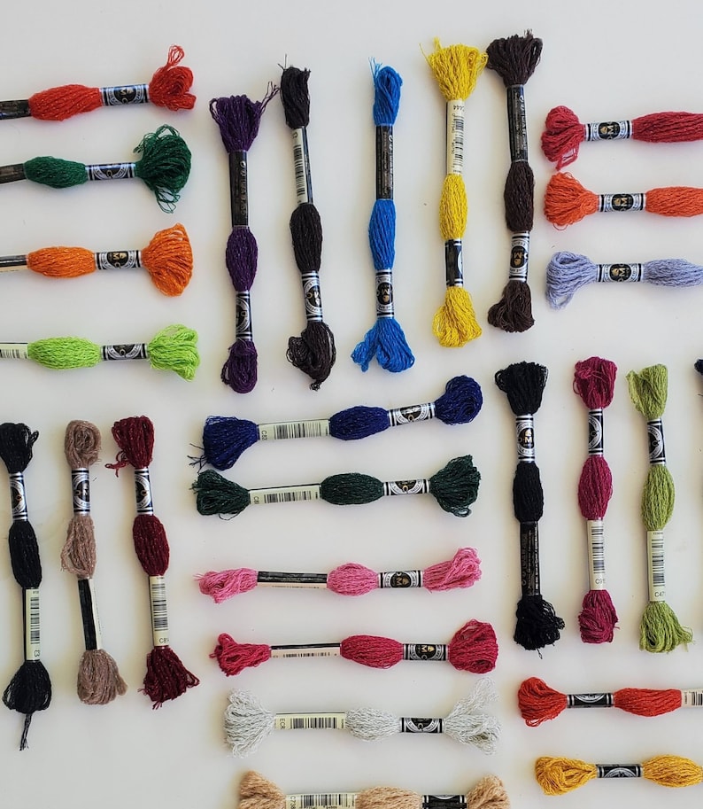 Puede incluir: Surtido de carretes de hilo de bordar en varios colores, incluyendo rojo, verde, azul, amarillo, morado, marr&oacute;n y gris. Los carretes est&aacute;n dispuestos en filas sobre una superficie blanca.