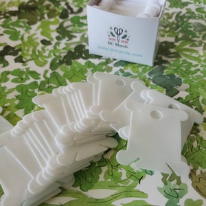 May include: A stack of white plastic thread bobbins, with a box labeled "BC Hands" and a website address. The bobbins are used for storing and organizing thread.