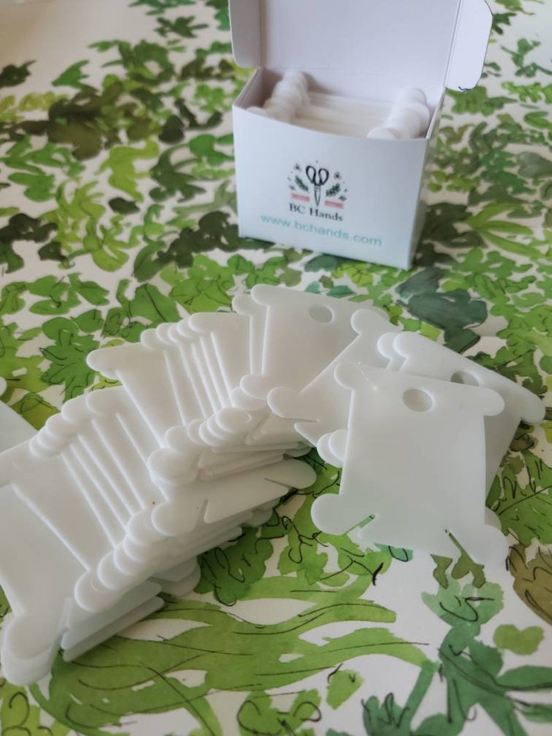 May include: A stack of white plastic thread bobbins, with a box labeled "BC Hands" and a website address. The bobbins are used for storing and organizing thread.