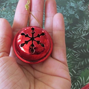 May include: A shiny red Christmas bell with a snowflake design. The bell is held in a hand and has a gold string for hanging. The background features a festive green and red pattern.