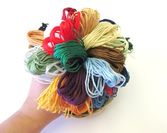 May include: A collection of colorful embroidery floss spools in a variety of shades, including red, green, blue, yellow, and brown. The spools are arranged in a circular pattern and are held together by a hand.